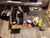 Overview photo showing the lockable aluminum case, cardboard box with chargers and cords, and various smaller items arranged on the floor