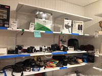 Shelves with multiple cameras, bags, flash, and boxes including visible Pentax, Olympus, Agfa cameras, and camera bags