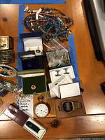 Overview image showing beaded necklaces, cuff links, pocket watches, Victorinox tool, pins, Calvin Klein nail file set spread out on wooden table.