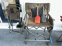 Two brown Cabela's folding chairs, one open with folding orange and grey shovels on the seat, the other chair folded. Both chairs have side trays with compartments.