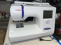 Front view of white Janome sewing machine showing the brand name, LCD screen, and stitching area.