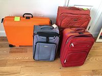 Four pieces of luggage lined up on wood floor by white wall: large orange Samsonite hard-shell, red quilted Ellen Tracy, red soft-sided, gray Samsonite soft-sided.