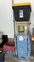 Stacked storage bins including a large Greenmade bin with yellow lid, beige RTIC cooler below, and several blue and one red plastic crates stacked beside them in a garage.