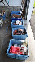 Five large blue plastic tubs positioned on a concrete floor, filled with various hand tools and hardware supplies visible from above and at angle.