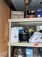 Shelving unit with various kitchen appliances and cookware including boxed Cuisinart food processor, cast iron pot, metal pots, and containers.