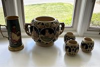 Wide shot showing the large container with handles, three matching mugs with castle reliefs, and the painted beer stein with a figure, all displayed on a window sill.