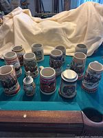Full view of the 13 beer steins arranged on teal surface against a beige backdrop showing variety of designs and sizes.