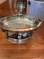 Oval stainless steel chafing dish on wooden floor, showing clear glass lid with metal handle, dual side handles, and metal base for fuel holder.