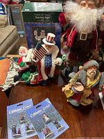 Photo showing Fabriche Musical American Santa and Uncle Sam Santa with American flag, seated Santa, and packaged Cobblestone Corner collectible ornaments.