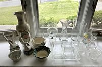 Various decorative glass and ceramic items displayed on a table by a window including a tall floral ceramic vase, two ceramic swan figurines, several clear glass vases of different shapes, and small ceramic bowls.