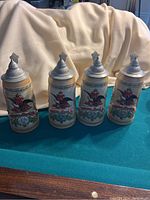 Front view of the four Anheuser-Busch ceramic steins showing the painted fronts and pewter lids with embossed logos