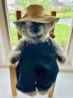 Front view of the jointed bear wearing a straw hat and navy pinstriped overalls, seated in a wooden chair by a window.