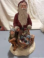 Front view of detailed painted resin Santa Claus figurine standing behind a sled filled with toys and accompanied by a small elf figure.