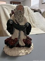 Full view of the Father Santa figurine showing cream and black robes, red poinsettias, snowy base, and gifts bag.