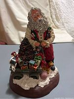 Front view of Santa Claus figurine holding wagon filled with gifts with Christmas tree and snow base visible.
