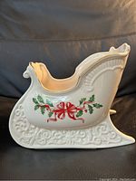 Left side view of the white porcelain sleigh with holly and red bow decoration, showing gold rim and embossed base.