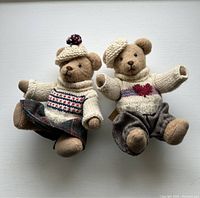 Photo showing both plush bears seated side by side, fully dressed in knitted sweaters, wool skirts, and cream knitted hats.