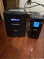 Two APC UPS units side by side on a hardwood floor, showing digital display screens and power cables, screens displaying status including 'Output OFF' and battery messages.