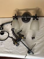 Two-light bathroom wall sconce with frosted glass shades and accompanying bathroom hardware items including faucet, towel ring, knob, and toilet paper holder on a white cloth.