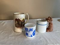 Four coffee mugs on a table including a ceramic cottage mug, a small Ranger Joe milk glass mug, a white mug with blue stripes, and a white mug with a 3D ET figure attached.
