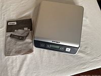 Digital postal scale with its user manual laid out on a fabric surface