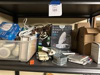 Shelf with various electrical supplies including LED bulbs in boxes, extension cords, boxes with electrical outlets and switches, and metal outlet box frame.