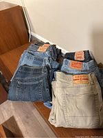 Top view of six pairs of men's jeans folded and stacked on a wooden surface, showing various washes and brand patches including Levi's, Lucky Brand, and Izod.