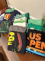 Photo showing folded men's t-shirts in multiple colors and various printed graphics, including a black shirt with a yellow and red Bob Marley face.