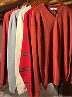 Five men's sweaters hanging on wooden hangers in various colors including brick red, grey, cream, and red with stripes.