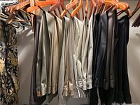 Photo of 14 pairs of men's trousers hanging on wooden hangers in various neutral colors including beige, brown, black, and khaki in a closet.