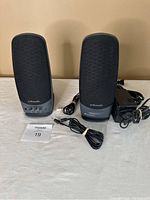 Two Polk Audio SGI speakers placed side by side on a white surface with cables and power adapter included.