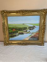 Frontal view of oil painting with Dutch landscape showing canal, boats, houses and green fields under blue sky in ornate gold frame