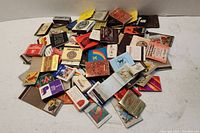 Photo showing a pile of approximately 100 assorted books of matches with varied colorful designs, logos and texts scattered on a white surface.