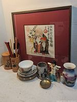 Photo showing entire lot with framed artwork in the background, wooden chopstick holders, ceramic cup, plates, small brass bowl, figurine, and vase.