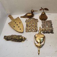 Top view of brass items including light switch covers, door knockers, figurines and decorative pieces on a white background.