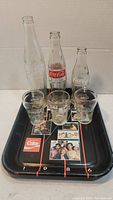 Three vintage Coca-Cola glass bottles, three Coca-Cola branded glasses, and a black metal Coca-Cola tin tray with 1981 graphics.