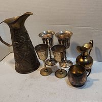 Seven pieces including large embossed copper pitcher, four etched goblets in two sizes, small brass pitcher, and small brass cup.