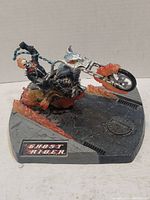Angled view of Ghost Rider figurine on motorcycle showing flaming front wheel, chain in hand, and base with Ghost Rider plaque and street texture.
