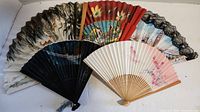 Photo of 5 decorative hand fans showing various floral and scenic designs, spread out to display patterns and colors including white, black, red, and cream bases.