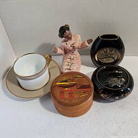 Overview of all five items together including figurine, tea cup and saucer, black dish and vase with gold edging, and carved wooden coaster set. Can for size reference.