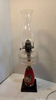 Full view of vintage oil lamp showing clear chimney, textured cut glass reservoir, and red base with floral art on black pedestal