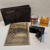 Photo of entire lot showing vintage photo album, Ansco film developer can, compact black camera, Kodakchrome II film, and 'Dear Old London' photographs pack