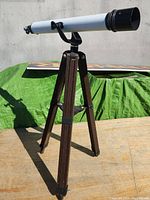 Full side view of the white telescope mounted on a wooden tripod on an outdoor surface with a green tarp in the background.