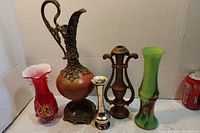 Photo showing five decorative vases including red glass, ornate metal ewer, vintage metal urn, silver-toned small vase, and tall green glass vase with a Coca-Cola can for scale.