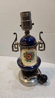 Front view of vintage blue ceramic lamp with floral design and metal decorative handles, showing socket and cord.