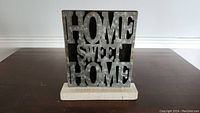 Full view of the galvanized metal 'HOME SWEET HOME' sign standing on a wooden block base against a white wall and dark floor.