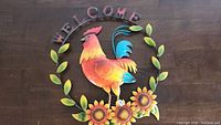 Front view of colorful metal rooster welcome sign laid on wooden surface.