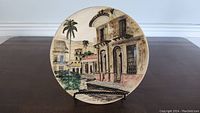 Front view of the 10-inch round decorative plate showing the painted scene of buildings, palm trees, and greenery with muted earth tones.