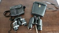 Two pairs of binoculars side by side on a wooden surface, each with its own carrying case.