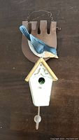 Front view of the decorative hanging bird feeder showing the full birdhouse, attached blue bird figure, rust-colored backing, and wire hanger.